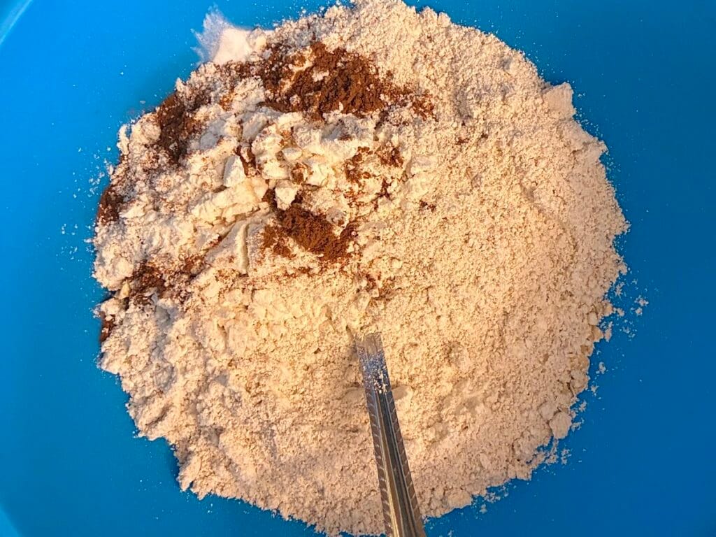 A stainless steel fork mixing quick-cooking oats, flour, baking soda, and warm cinnamon together in a large blue mixing bowl.