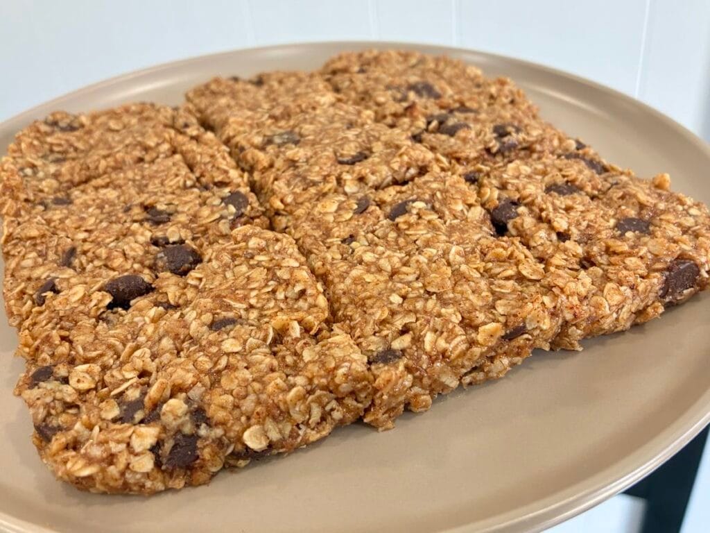 Fudgy Almond Butter No-Bake Cookie Bars 3 Angled shot of whole, un-cut fudgy almond butter no-bake cookie bars resting on a clean matte stoneware plate, with dark chocolate pockets visible across the top.