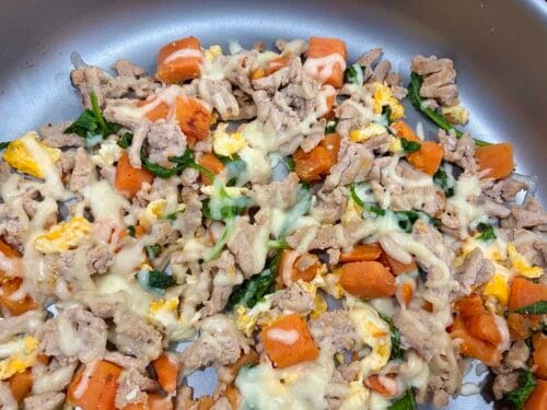 Overhead view of a high-protein turkey and sweet potato breakfast hash melt inside a medium-large skillet, topped with a bubbling layer of melted mozzarella cheese and wilted spinach.
