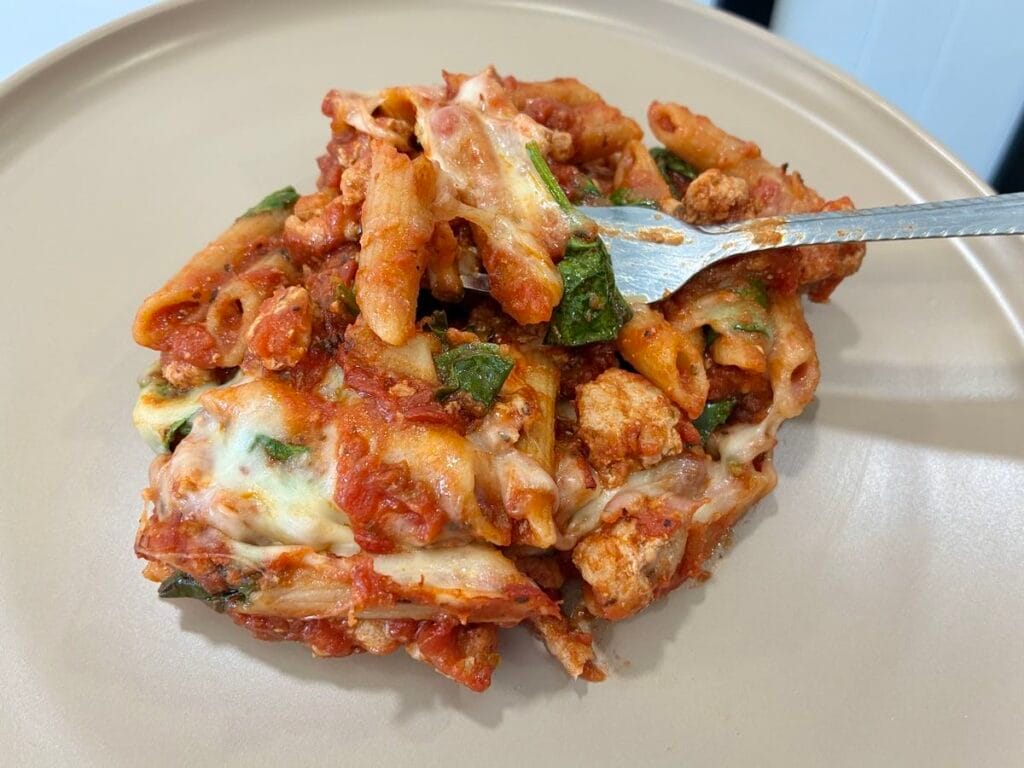 A generous serving of healthy turkey baked ziti with wilted spinach and melted cheese on a round matte stoneware dinner plate.