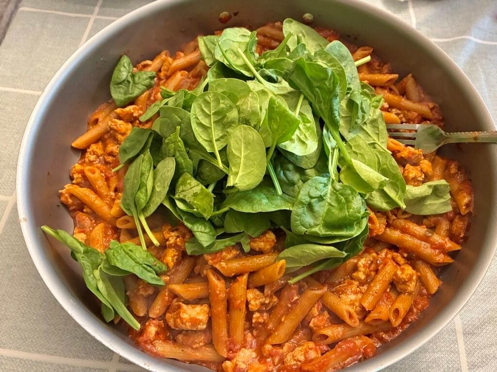 A mountain of fresh green baby spinach leaves piled high on top of ground turkey pasta inside a cooking skillet.
