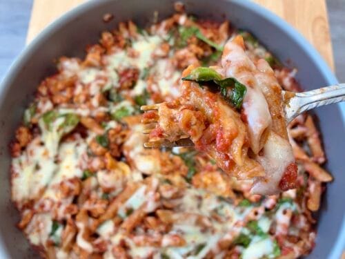 A close-up macro shot of a fork lifting a bite of cheesy turkey baked ziti with spinach leaves, with the cooking skillet softly blurred in the bright background natural light.