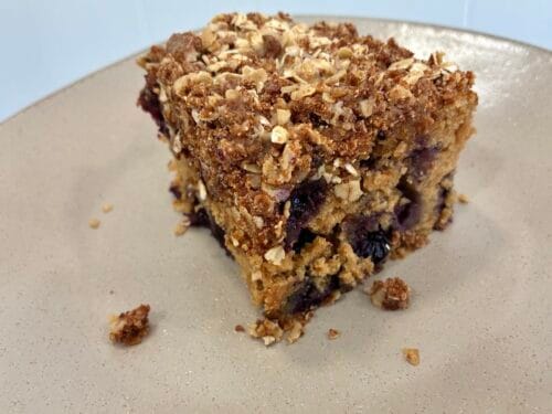 A bright, high-contrast food photography shot taken in the natural light of an outdoor breezeway, showing a single square slice of one-bowl blueberry cinnamon coffee cake served on a plate. The moist cake crumb is loaded with fresh blueberries and topped with a thick, rustic layer of crisp oat cinnamon streusel crumbs.