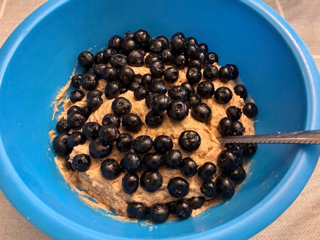A close-up recipe process shot looking down into a mixing bowl, showing a thick layer of vibrant, fresh blue blueberries resting on top of a thick, creamy coffee cake batter right before being gently folded into the mixture.