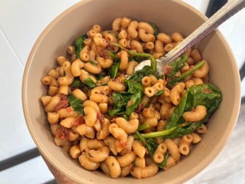 A close-up process shot of hot pasta simmering inside a saucepan, showing a stainless steel fork gently folding fresh baby spinach leaves into a vibrant red sun-dried tomato cream sauce as the greens wilt from the ambient heat.