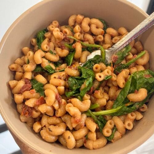 A close-up process shot of hot pasta simmering inside a saucepan, showing a stainless steel fork gently folding fresh baby spinach leaves into a vibrant red sun-dried tomato cream sauce as the greens wilt from the ambient heat.