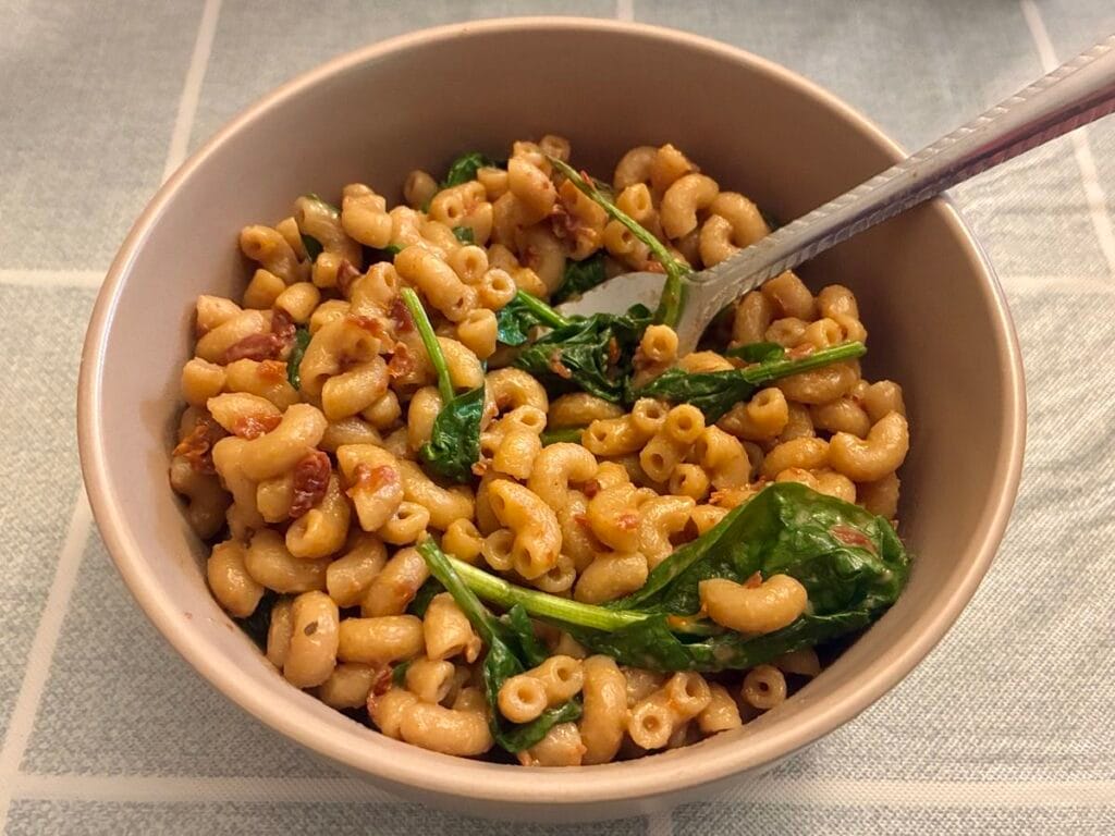 A cozy lifestyle photography shot showing a finished portion of healthy sun-dried tomato and spinach pasta served inside a gorgeous matte stoneware bowl sitting on a living room table, evoking a warm, comforting home dinner vibe.