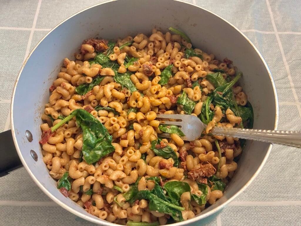 An overhead studio food photography shot of a 15-minute one-pot creamy sun-dried tomato and spinach pasta served warm in a matte stoneware bowl out on a sunlit breezeway. Organic macaroni noodles are coated in a rich, dairy-free tomato cream sauce, mixed with deep green wilted baby spinach and pieces of bright red sun-dried tomatoes.