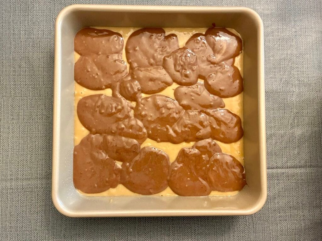 Overhead view of chocolate cake batter dollops dropped on top of a smooth vanilla cake batter base in a square baking pan.