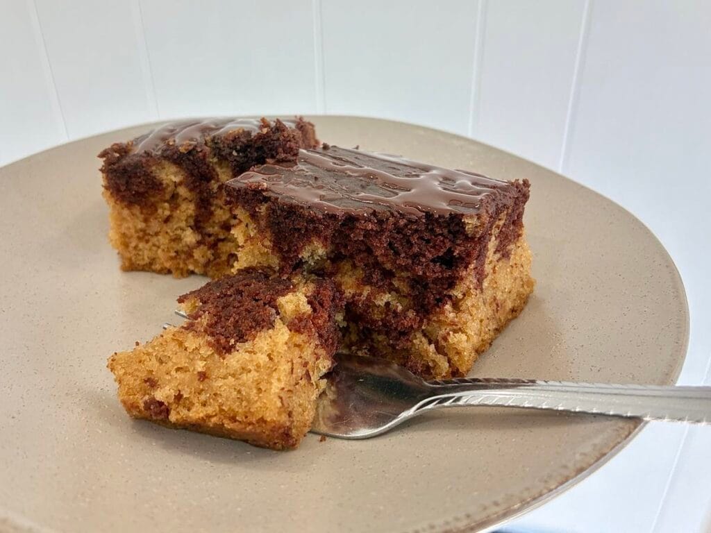 A close up look at slices of eggless marble snack cake, topped with a chocolate glaze; on a plate with a dessert fork breaking off a bite to show the soft interior crumb.