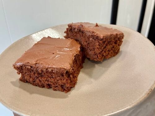 Two decadent square slices of gluten-free oat flour chocolate cake topped with a thick layer of homemade chocolate frosting, served on a clean dessert plate.