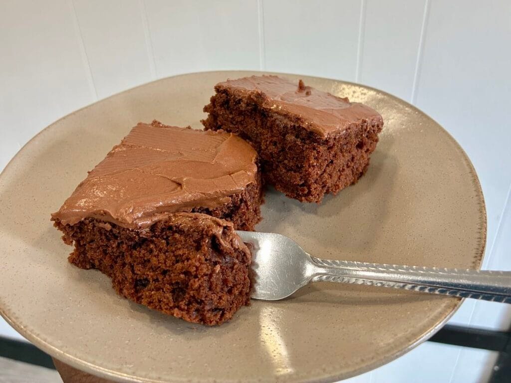 A dessert fork breaking off a bite of ultra-moist oat flour chocolate cake, showing the texture of the homemade chocolate frosting and the soft cake layer underneath.