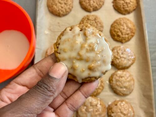 A close-up view of a hand holding up one of the thick, soft chewy iced oatmeal cookies toward the camera, with the rest of the glazed cookies on a baking sheet, and a bowl of glaze in the background.