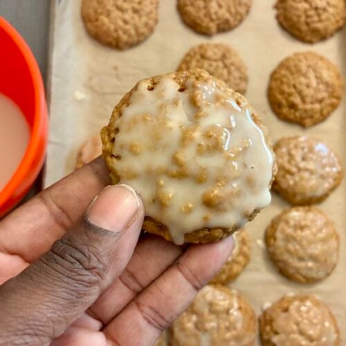 A close-up view of a hand holding up one of the thick, soft chewy iced oatmeal cookies toward the camera, with the rest of the glazed cookies on a baking sheet, and a bowl of glaze in the background.