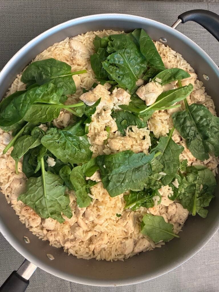 Fresh baby spinach scattered over a skillet of creamy garlic chicken and rice before fluffing.
