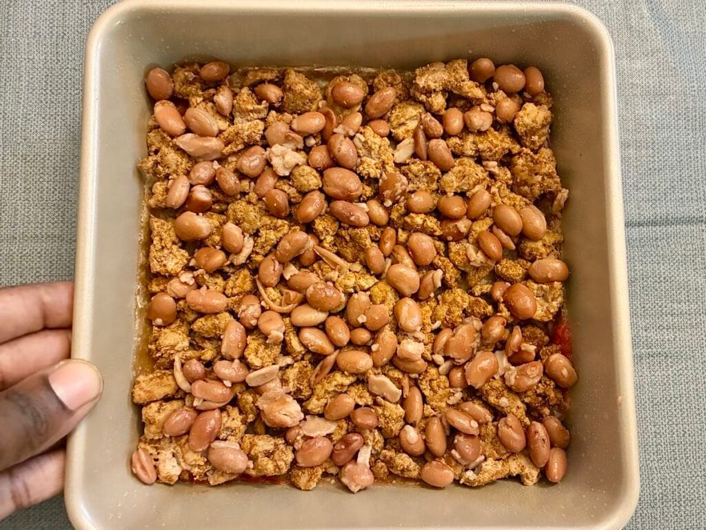 Assembling the first layers of healthy taco casserole with tomato sauce, a flour tortilla, seasoned meat mixture, and pinto beans in an eight by eight inch pan.