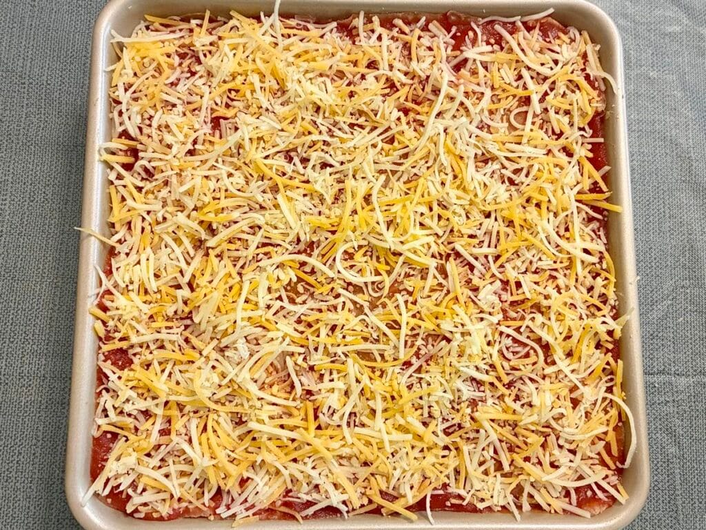 The completed layers of the healthy taco casserole topped with a generous layer of shredded Mexican cheese in an eight by eight baking pan before going into the oven.