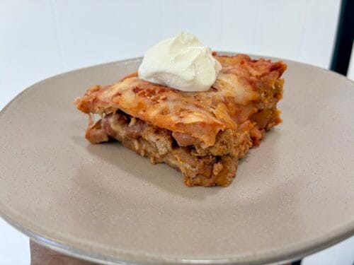 A square slice of healthy layered taco casserole served on a white plate and topped with a dollop of sour cream.