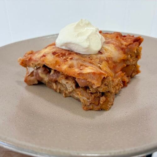 A square slice of healthy layered taco casserole served on a white plate and topped with a dollop of sour cream.