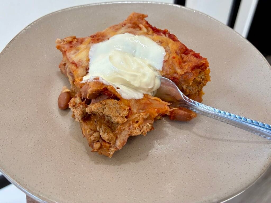 A close-up view of a fork cutting into a square slice of healthy taco casserole with a dollop of sour cream on a plate, showing the soft, delicious layers.