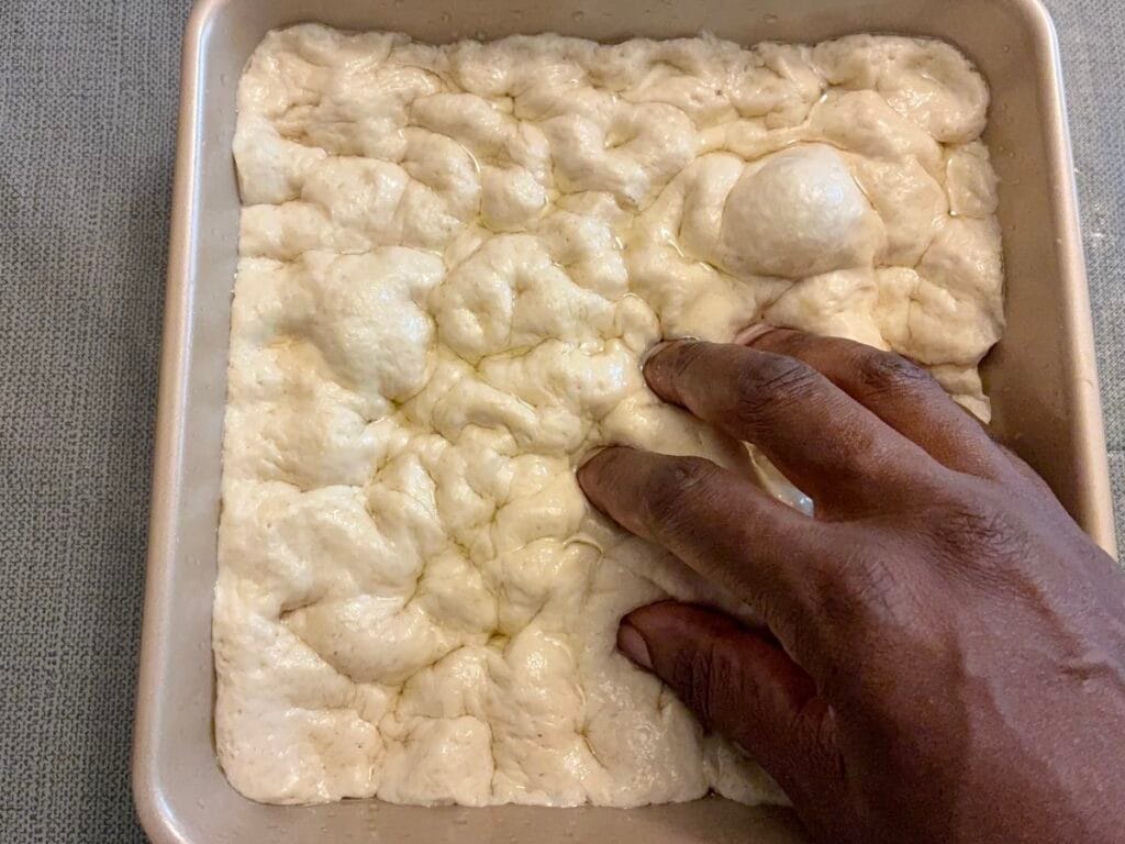 Easy 8x8 No-Knead Focaccia (Beginner Friendly) 4 Oiled fingers pressing deep dimples into puffy no knead focaccia dough inside an 8x8 square baking pan.