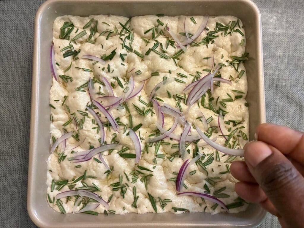 Hand scattering paper-thin red onion slices and fresh organic rosemary needles over dimpled no knead focaccia dough.