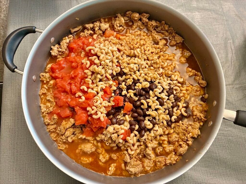 Diced tomatoes, black beans, raw elbow macaroni, and chicken broth added inside of a 12-inch skillet before cooking.