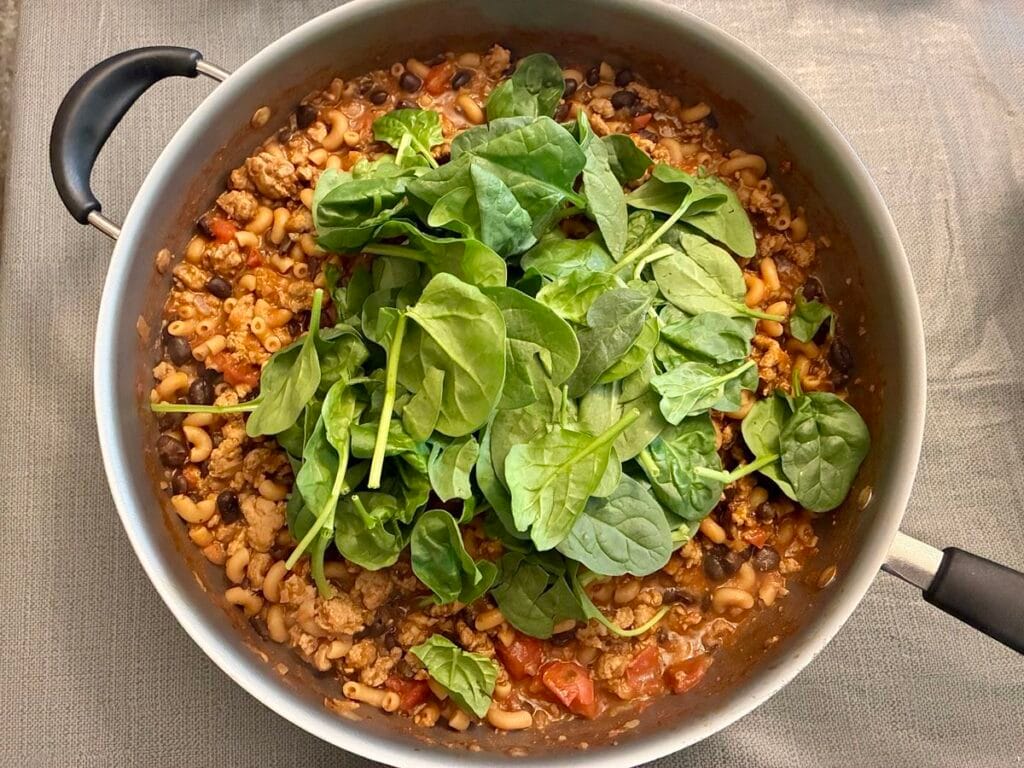 Fresh organic baby spinach leaves layered on top of an unmixed skillet of cooked ground turkey, black beans, diced tomatoes and elbow macaroni.