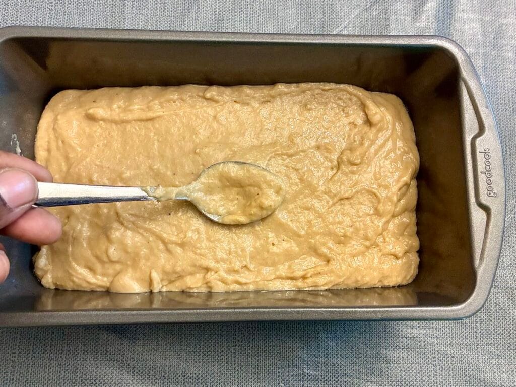 A small stainless steel spoon smoothing down the top of thick golden cake batter inside a 9x5-inch loaf pan.