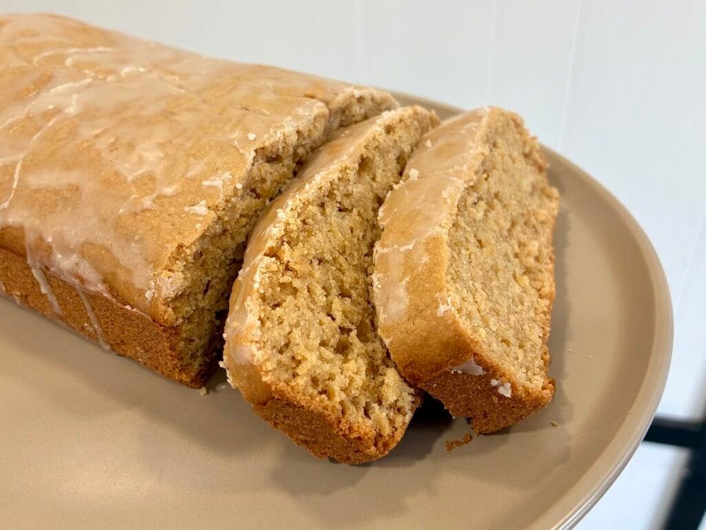 A whole glazed lemon cardamom loaf resting on a matte stoneware plate with a couple of cut slices staggered in front.