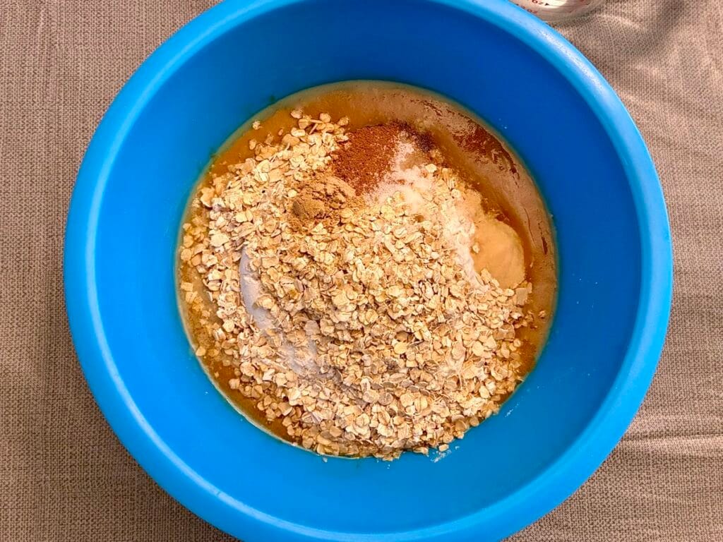 Healthy Eggless Pumpkin Chocolate Chip Snack Cake (8x8 Pan) 3 A top-down view of all the measured recipe ingredients inside a large blue mixing bowl, showing quick oats, flour, and dry spices layered over the wet mixture for the healthy eggless pumpkin chocolate chip snack cake.